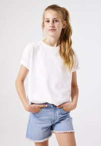 T-shirt blanc à manches courtes avec des poignets retroussés, associé à un short en denim bleu clair avec des ourlets effilochés. Le modèle pose les mains dans les poches.