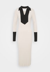 Abito in maglia a costine con colletto nero e scollatura a V, con un pannello nero a contrasto. Maniche lunghe e silhouette aderente. Colore: crema e nero.