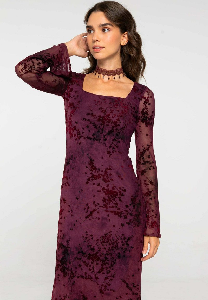 Vestido de terciopelo burdeos con patrón floral, escote cuadrado y mangas largas de tela transparente. Acentuado con un collar de cuentas alrededor del cuello.