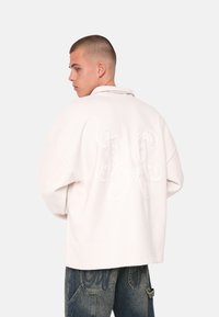 Lys rosa oversized jakke med en foldet krave og stort broderet design på ryggen. Blød tekstur, lange ærmer og afslappet pasform.