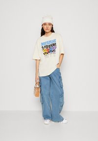 Överdimensionerad krämfärgad T-shirt med färgglada tecknade grafiker; blå baggy cargobyxor; vita sneakers; genomskinlig handväska; krämfärgad mössa.
