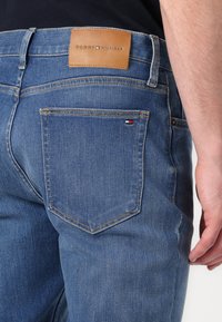 Plave traperice s kožnom etiketom na kojoj piše "TOMMY HILFIGER." Stražnji džep prikazuje mali logotip zastave, suptilan detalj šivanja.