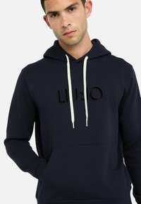 Un jeune homme portant un sweat à capuche bleu marine avec des cordons blancs, une poche frontale et un texte en relief noir sur la poitrine, se tenant devant un fond blanc.