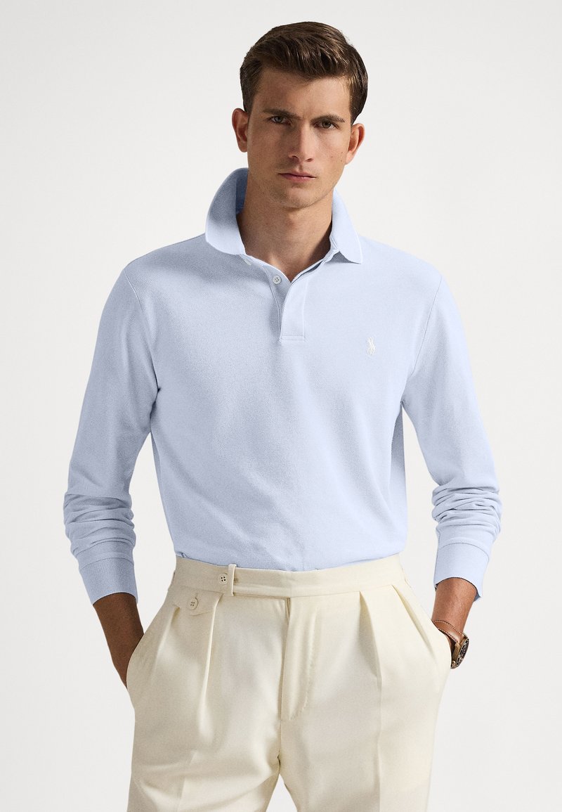 Άνδρας με κοντά καστανά μαλλιά φοράει ανοιχτό μπ�λε πουκάμισο πόλο με μακριά μανίκια, μπαίνει μέσα σε κρεμ παντελόνι ψηλόμεσο, έχοντας τα χέρια στις τσέπες.