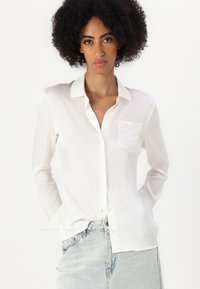Camisa blanca de manga larga con botón y cuello, que presenta un bolsillo en el pecho y una textura sutil, combinada con jeans de mezclilla claros.