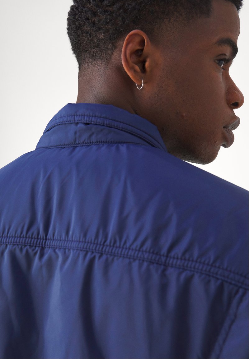 Chaqueta ligera azul marino con cuello alzado, costuras visibles y textura suave. Presenta un corte ajustado y un brillo sutil.