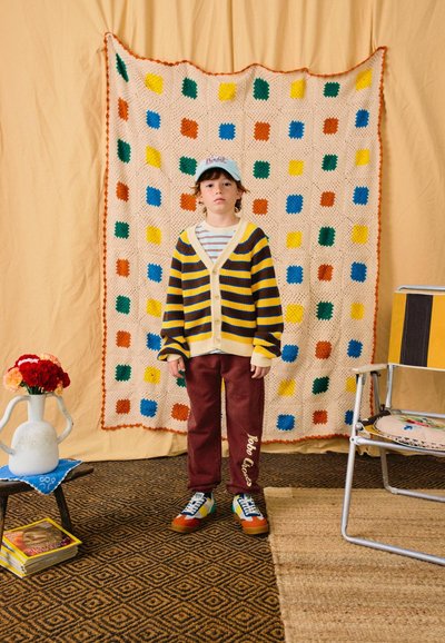 Dreng iført stribet cardigan, burgunderfarvede bukser og kasket står foran et flerfarvet hæklet tæppe hængende på en beige væg.