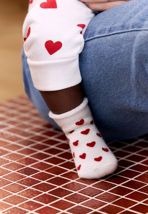Beebi, kes kannab valgeid sokke ja punaste südamekujude mustriga pükse, toetab jalga punaste plaatidega põrandal, kõrval inimene sinistes teksapükstes.