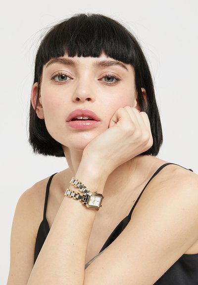 Jeune femme aux cheveux courts noirs portant un haut noir sans manches, reposant son menton sur sa main, avec une montre-bracelet en or et argent.