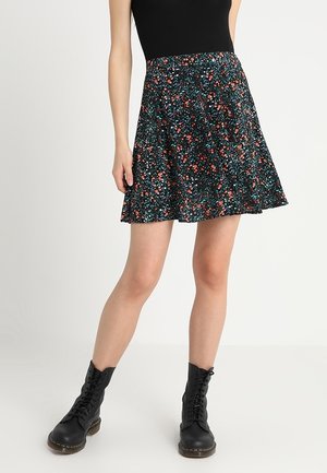 Frau trägt einen schwarzen A-Linien-Rock mit roten und türkisfarbenen Blumen, schwarzes ärmelloses Oberteil und schwarze Schnürstiefeletten.