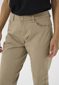 Pantalons en mélange de coton beige avec un design classique à cinq poches, coutures visibles et fermeture à bouton à l'avant. Texture lisse et style polyvalent.