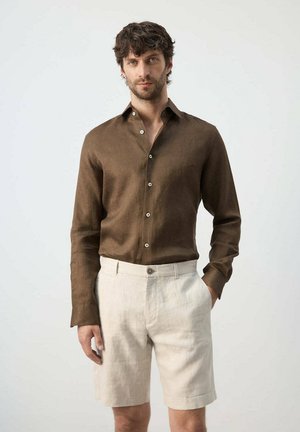Homme aux cheveux foncés portant une chemise marron à manches longues boutonnée et un short beige clair, debout avec une main dans la poche.