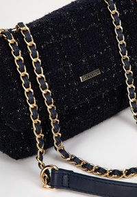 Marineblaue, strukturierte Handtasche mit einer goldenen Kettenriemen, die ein geflochtenes Design und eine markierte Metallplatte aufweist. Akzente setzen ein Trageriemen aus Leder.