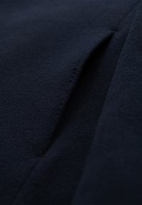 Dunkel marineblauer Wollstoff mit einer schmalen, vertikalen Taschenöffnung, die Textur und weiche Materialfalten zeigt.