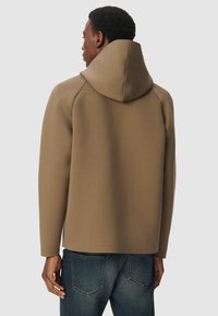 Mężczyzna w beżu bluzie z kapturem, odwrócony tyłem, w połączeniu z wyblakłymi niebieskimi dżinsami na jednolitym jasnym tle.