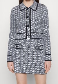 Kobieta w pasiaku w kolorze granatowym i białym, nosząca dopasowaną marynarkę i mini spódniczkę z czarnymi wykończeniami oraz dużymi granatowymi guzikami.