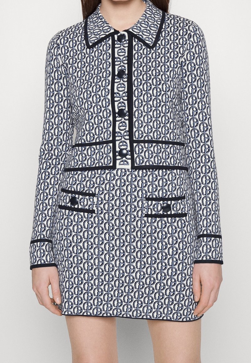 Kobieta w pasiaku w kolorze granatowym i białym, nosząca dopasowaną marynarkę i mini spódniczkę z czarnymi wykończeniami oraz dużymi granatowymi guzikami.