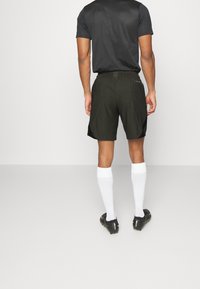 Uomo che indossa pantaloni sportivi neri, maglietta nera, calze bianche fino al ginocchio e scarpe cleats nere, in piedi contro uno sfondo bianco uniforme.