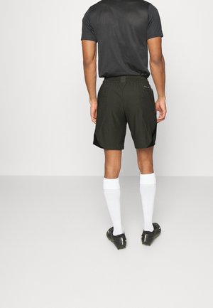 Homme portant un short de sport noir, un t-shirt noir, des chaussettes blanches jusqu'aux genoux et des chaussures à crampons noires, se tenant contre un fond blanc uni.