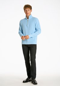 Ljust blå zip-up tröja med ribbad textur, ihopparad med svarta byxor och blanka svarta skor. Modellen står mot en vit bakgrund.