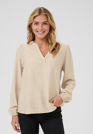 Chemisier beige à manches longues avec un col en V, des poignets élastiques et une coupe décontractée. Texture de tissu lisse et design simple, associé à un pantalon noir.