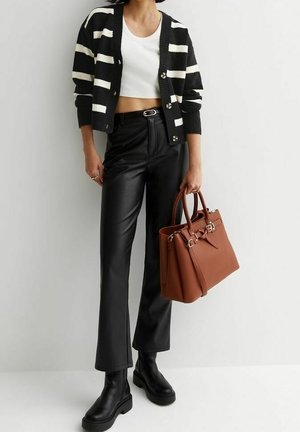 Vrouw met een zwart-wit gestreepte cardigan, een wit crop top, zwarte leren broek, zwarte laarzen, en een bruine leren handtas in haar hand.