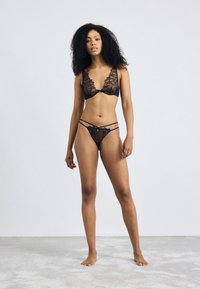 Ensemble de lingerie en dentelle noire comprenant un bralette en V profond avec broderie florale et un string assorti avec des fines bretelles. Texture douce, design délicat.