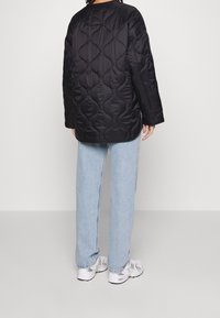 Černá prošívaná bunda s kulatým výstřihem a volným střihem, v kombinaci se světle modrými džínami a bílými teniskami. Hladká, lesklá textura.