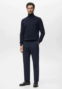 Jersey de cuello alto azul marino con una textura suave, combinado con pantalones oscuros de corte ajustado en azul marino y zapatos de cuero negros. El modelo está de pie con una expresión neutral.