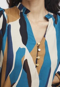 Blusa multicolore con motivi astratti blu, marrone e crema. Scollo a V con una placket di bottoni e dettagli sulle spalle plissettati.