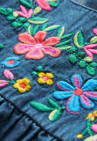 Fleurs brodées colorées et feuilles vertes sur tissu en denim bleu, avec des motifs floraux roses, jaunes et bleus.