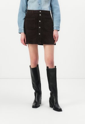 Kvinde iført en mørkebrun mini-nederdel i fløjl med knapper foran, lyseblå denimjakke og sorte knæhøje støvler med hæl mod hvid baggrund.