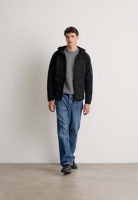 Jonge man die naar voren loopt, gekleed in een zwart hoodiejack, grijze trui, blauwe spijkerbroek en zwarte schoenen, tegen een effen witte achtergrond.