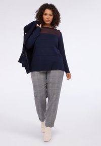 Jersey de marina con panel transparente, combinado con pantalones de cuadros gris claro. La modelo lleva botines crema y sostiene un blazer en una mano.