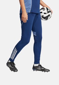 Marinefarbene Sportleggings mit einer figurbetonten Passform, ausgestattet mit weißen Akzentstreifen und einer Seitentasche. Getragen mit schwarzen Fußballschuhen und hält einen Fußball.