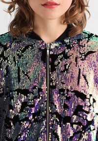 Veste bomber scintillante ornée de sequins multicolores sur un fond noir, fermeture éclair et col côtelé, présentant une surface texturée.