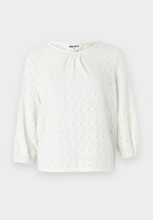 Hvit bluse med rund halsutsnitt og trekvartlange pufferm, med et teksturert vevd mønster og subtile diamantformede utskjæringer.