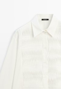 Chemise blanche avec un design structuré comprenant des plis horizontaux sur le devant, un col classique et une fermeture à boutons.