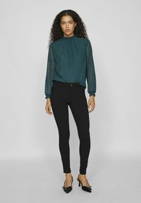 Blusa a fantasia turchese con collo alto e maniche lunghe, abbinata a jeans skinny neri e scarpe con tacco nere.