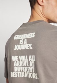 Camiseta de algodón gris con un gran texto blanco en la parte de atrás que dice "LA GRANDEZA ES UN VIAJE. TODOS LLEGAREMOS A DESTINOS DIFERENTES."