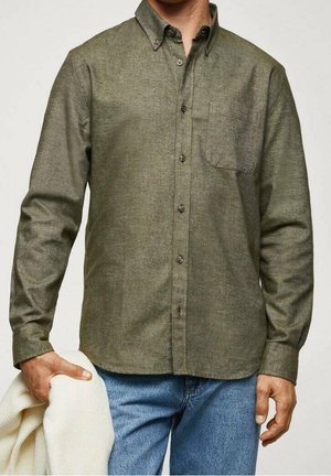 Man draagt een olijfgroene overhemd met knopen en een blauwe spijkerbroek, houdt een opgevouwen witte trui in zijn linkerhand.