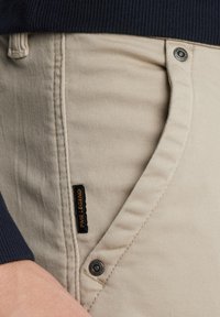 Beige chinos med en glat tekstur, der har en sidelomme, knaplukning og et sort mærke med "PINE LEGEND" på taljen.