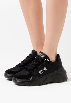 Sneaker nere che presentano una combinazione di materiali lisci e testurizzati, suola spessa e il marchio "VERSACE JEANS COUTURE" sulla linguetta e sul lato.