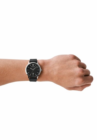 Handgelenk mit einer schwarzen Lederarmbanduhr mit schwarzem rundem Zifferblatt, silbernen Markierungen und einer Datumsanzeige bei 6 Uhr vor weißem Hintergrund.