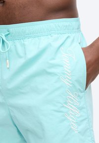 Κοντινό πλάνο ενός ατόμου που φοράει ανοιχτό μπλε μαγιό με την επιγραφή "Tommy Hilfiger" κεντημένη κάθετα δίπλα στην πλαϊνή τσέπη.