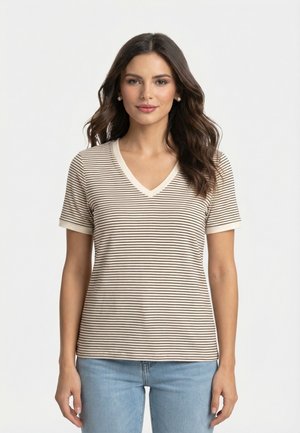 Femme aux longs cheveux ondulés portant des boucles d'oreilles en perles, un haut beige et noir à rayures avec col en V à manches courtes, et un jean bleu clair, debout devant un fond blanc.