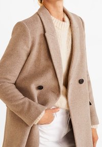 Femme portant un manteau beige à double boutonnage sur un pull en tricot crème avec un pantalon blanc, main dans la poche, debout contre un fond blanc.