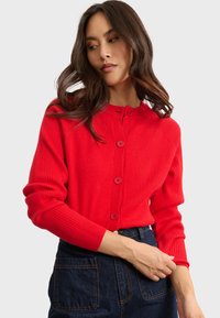Cardigan rosso a coste con scollatura rotonda, cinque bottoni e maniche lunghe. Indossato sopra jeans scuri, evidenziando una silhouette aderente.
