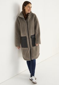 Frau, die an einer Wand lehnt und einen langen hellbraunen Fleece-Mantel mit schwarzen Taschen, dunklen Jeans und weißen Sneakers mit schwarzen Streifen trägt.
