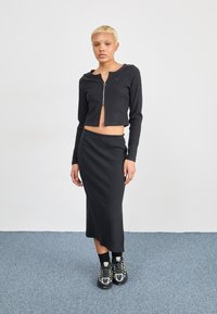 Svart ribbestrikket langermet topp med glidelås og matchende knelangt skjørt. Modellen har på seg svarte joggesko med neonlys og svarte sokker.